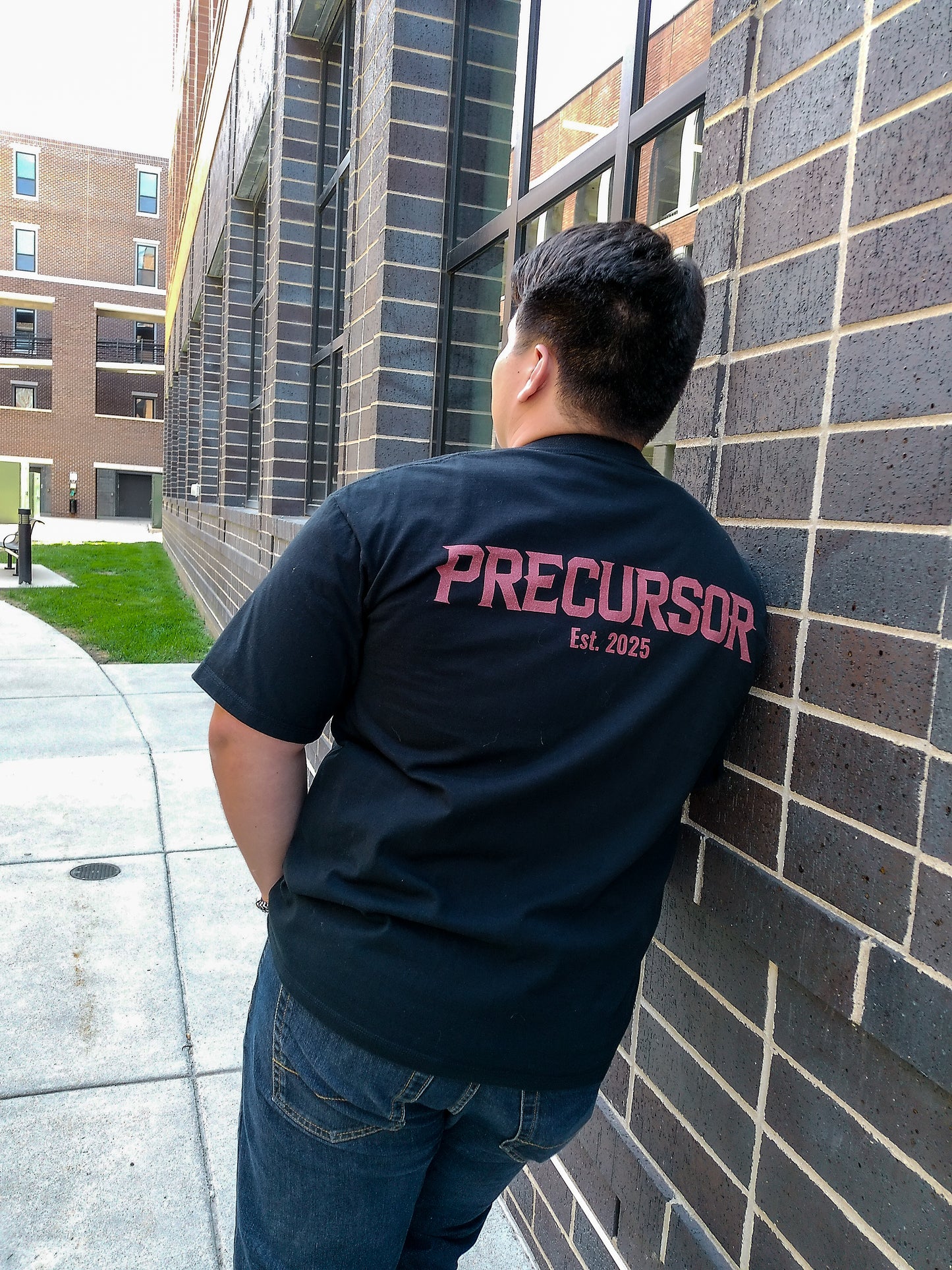 Person wearing a black t-shirt with 'PRECEDOR EST. 2023' text, leaning against a brick wall.