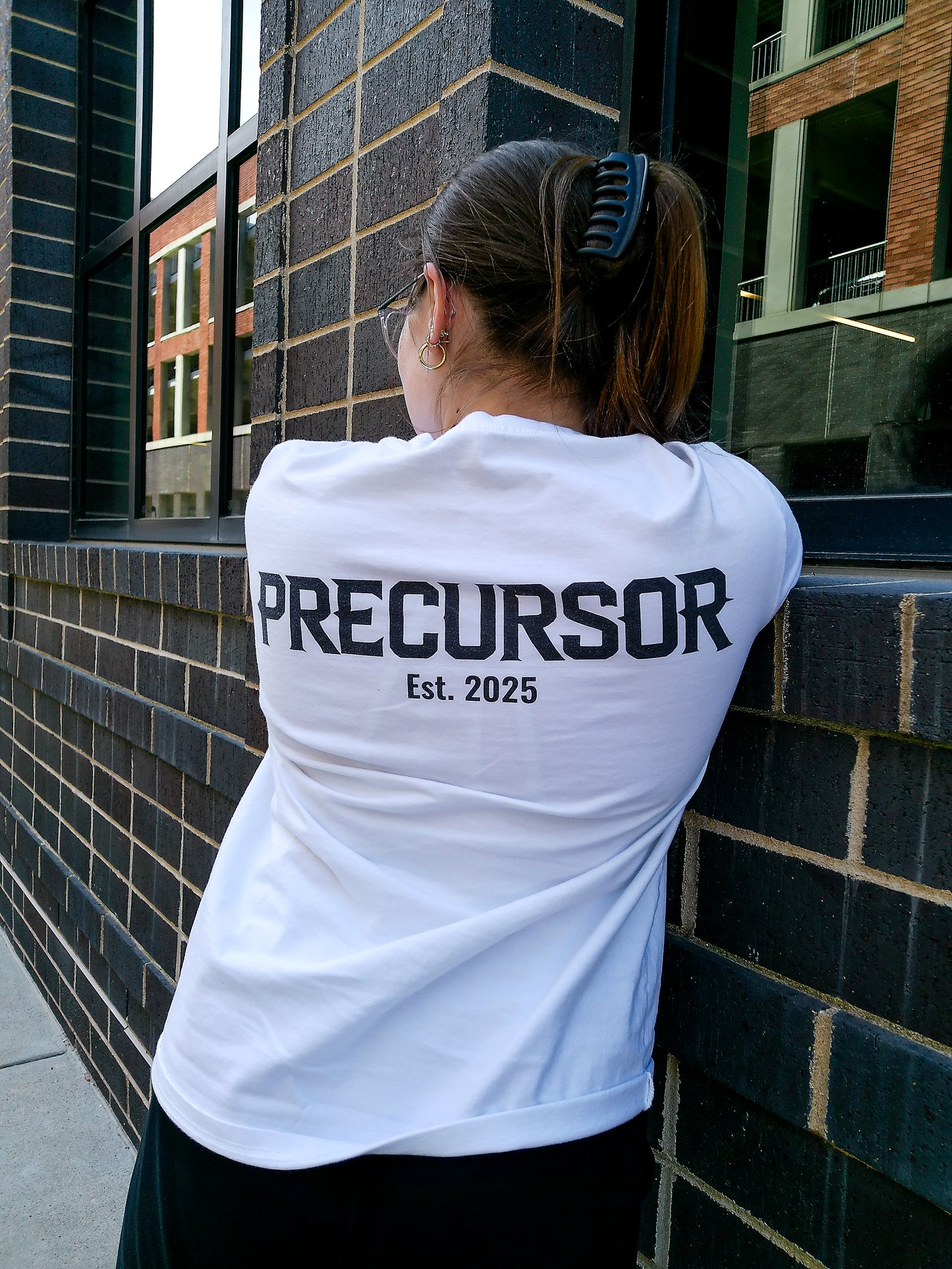 Person wearing a white t-shirt with 'PRECURSOR Est. 2025' text, leaning against a brick wall.