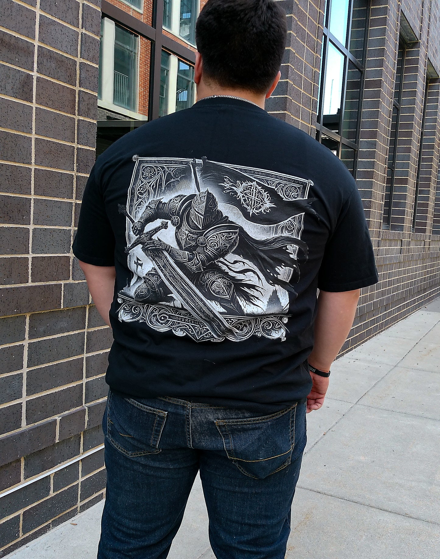 Person wearing a black t-shirt with a graphic design on the back, standing against a brick wall.