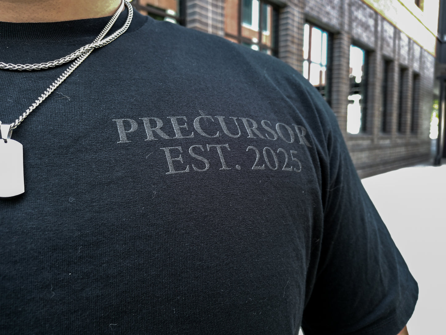 Person wearing a black shirt with 'PRECURSOR EST. 2025' text, standing outdoors.